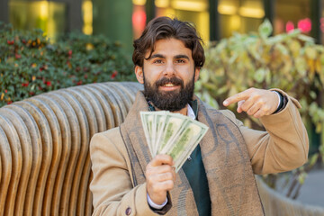 Successful rich young Indian man holding and waving dollar bill money fan on downtown city street. Happy adult Hindu guy winning online lottery game offers a cash loan for purchases calculating budget