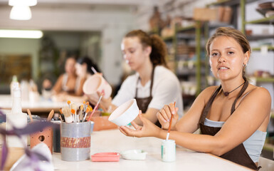 Interested young woman in pinafore drawing on handmade clay jar with paintbrush in art studio