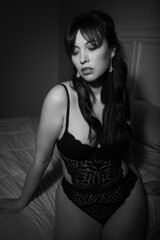 Black and white portrait of a 30-year-old Canadian caucasian woman wearing a black lace bodysuit, sitting on her bed in the bedroom and looking sultry at the camera. Intimate and moody.