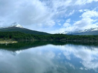 Stunning landscape of a serene alpine lake with crystal-clear reflections of snow-capped mountains and lush forest under a dynamic sky. Captured in pristine Patagonia.