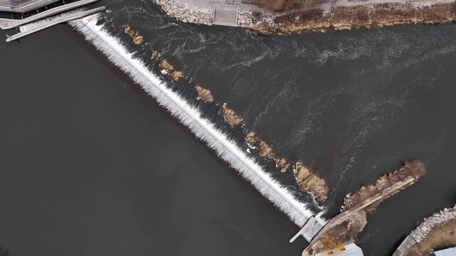 Drone footage of a small dam in a tranquil natural setting. The water flows gently over the structure, surrounded by lush greenery and rocky terrain, creating a serene and peaceful scene.
