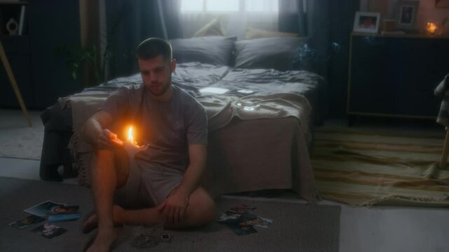 Full low key shot of young heartbroken and melancholic man sitting on floor near bed among pictures and burning photo of ex-wife, feeling deep pain and sorrow