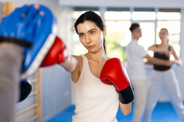 Diligent young lady wearing boxing gloves practicing punches with trainer during sports classes