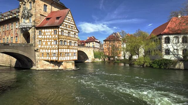 A well-lit Bright sunny day for tourism with blue sky and flowing water in the foreground and beautiful architecture and tourists on a bridge in the background in Bamberg Bayern Bavaria Germany Europe