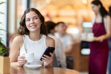 Young woman is sitting at a table in a cafe, drinking hot strong coffee from natural beans and holding a mobile phone in hands. Coffee shop visitor checking social networks on her phone