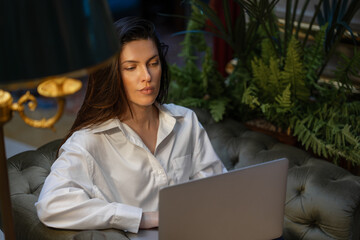 Businesswoman using laptop while sitting on cushioned chair in hotel lobby. Young woman in white shirt and appears focused on work. Plants and warm lighting create calm, professional atmosphere