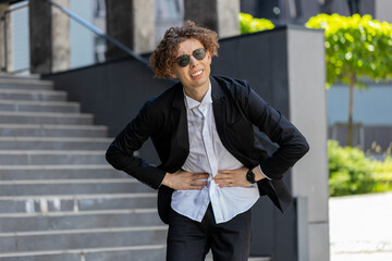 Young Caucasian businessman in formal suit feels stomach pain standing on the street near office...