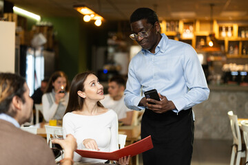 In dim light of restaurant hall, African male waiter communicates with girl customer, answers questions about composition of dish of day, serve couple in restaurant