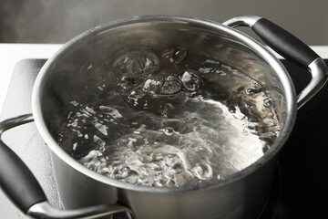 Cooking pot with boiling water and stove on white table against grey background, closeup