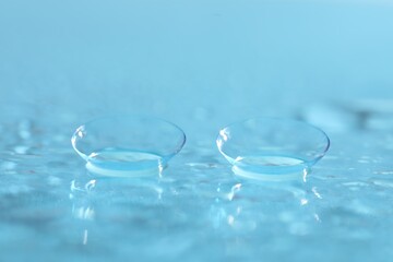 Pair of contact lenses and water drops on light blue mirror surface, closeup
