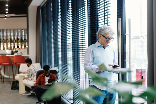 Mature IT company boss using smartphone near window near employees