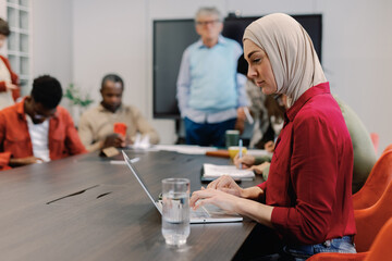 Young muslim female digital developer typing on laptop near colleagues