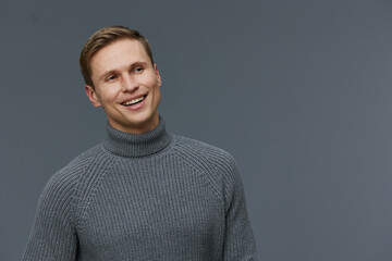 Fototapeta premium Smiling young man in gray turtleneck sweater looking happy against plain gray background, studio portrait, casual style, cheerful mood, modern fashion, male model, warm expression, studio photography