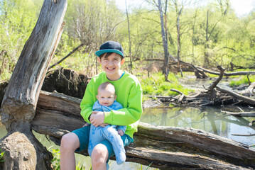happy 12 year old boy holding his 4 month old baby brother, sitting on log by river in spring	