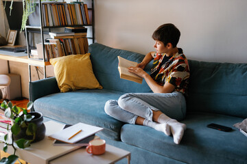 Young concentrated woman reading book on couch at home at daytime