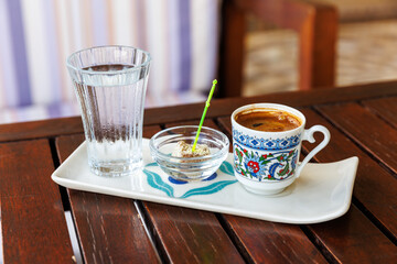 Turkish coffee served in ornate cup with glass of water and piece of Turkish delight on tray, set on dark wooden table, cultural tradition.