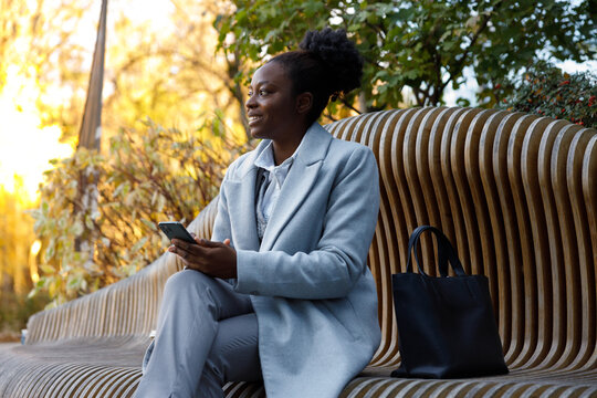 Woman in formal wear sitting on bench in park with smartphone in hands