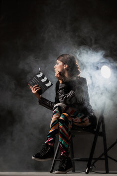 Female movie director on set with smoke background. Girl holding clapperboard on fire.