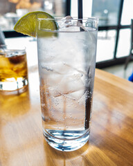 A crisp gin and tonic served in a tall glass packed with ice, topped with a lime wedge. Tiny bubbles rise through the clear drink, catching the sunlight on a wooden table by a bright window.
