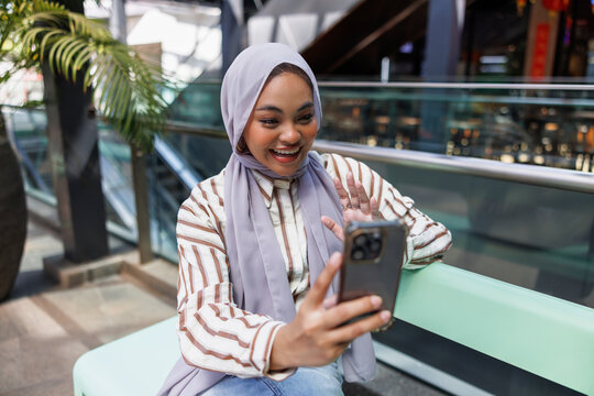 Cheerful woman in hijab video calling through smartphone on bench