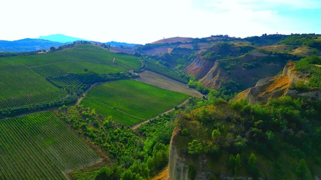 Sumptuous aerial pullback pan left shot from Offida, a view near Chiesa di Santa Maria della Rocca with gorgeous striped green fields, steep vegetation-filled promontories, hillscapes, blue mountains