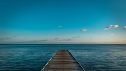 Obraz premium A tranquil photograph featuring a long wooden pier extending straight out over a calm, deep blue ocean towards the horizon.