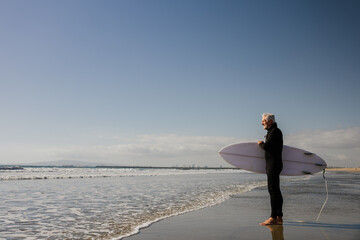 Elderly Surfer Standing on Beach With Surfboard on a Clear Day