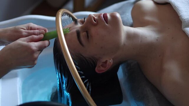 Close-up of a woman receiving a relaxing Japanese head spa treatment, with green massage sticks applied to her scalp, while water gently flows from a special tool during the session in a beauty salon