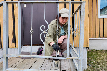 Middle aged woman paints metal railing of porch with black paint using paintbrush, paint can placed...