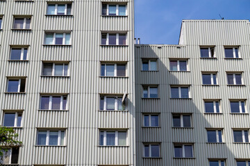 the facade of a multi-storey residential building covered with vertical light grey facade panels. The building has numerous windows, some of which are open. On the right side, you can see the shadow o