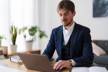 A young professional man in a suit works on his laptop at a desk. He is focused on his work in a bright, modern office space.