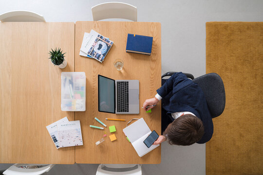 Overhead shot of a businessman working at his desk. He is using a smartphone and taking notes in a notebook while working on his laptop.