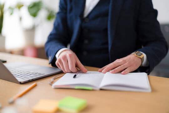 A businessman reviews notes in a notebook at his desk. He is wearing a suit jacket and has a laptop nearby.