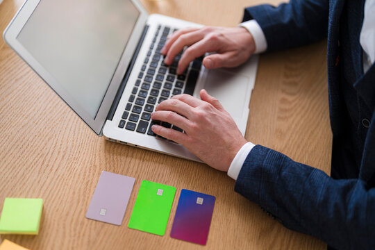 A person types on a laptop with multiple credit cards nearby. He is wearing a suit jacket and working at a desk.