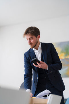 A businessman in a suit checks his phone while reviewing documents at his desk. He is focused on his work and staying connected.