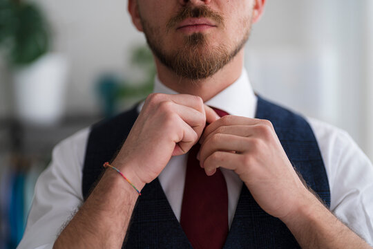 A bearded man in a vest adjusts his red tie. He is wearing a white shirt and has a colorful bracelet on his wrist.