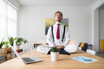 A businessman meditates on a desk in a bright office. He is wearing a shirt, tie, and suspenders, finding calm amidst the workday.