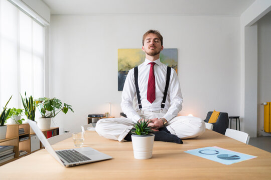 A businessman meditates on a desk in a bright office. He is wearing a shirt, tie, and suspenders, finding calm amidst the workday.