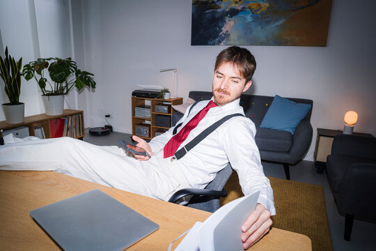 A man in a shirt, tie, and suspenders relaxes at his desk. He's using his phone and has a laptop and router nearby in a modern, comfortable home office.