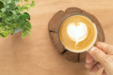Hand hold the cappuccino displays latte art in the form of a heart, created with white foam against the light brown coffee on a brown mug over a circular slice of wood over wood background. Top view..