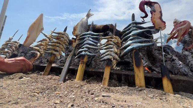 Skilled chef grilling fresh seafood skewers on sandy Mediterranean beach, positioning octopus and fish over glowing charcoal embers. Typical "espetos" food of Spain, Costa del Sol, Malaga.