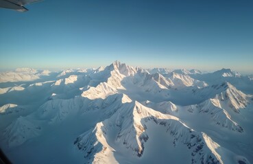 Aerial view of snowy mountain peaks under clear blue sky. Flight over winter landscape. Cold glacier, snow covered rocks, ice. High altitude view from the airplane window. Mountain range travel tour.