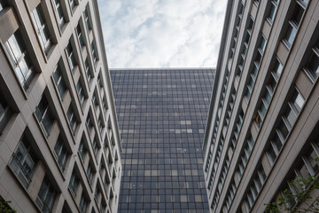 Upward view between two modern office buildings revealing a tall glass skyscraper against a lightly clouded sky, showcasing urban density and geometric architectural lines in a corporate setting.

