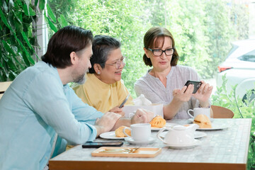 Senior Asian man and two Caucasian adults laugh together at a cafe table during casual conversation. The cheerful elderly group enjoys spending free time recreation or retire community in bakery shop