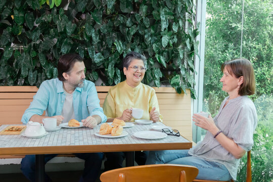 Caucasian man and senior Asian man chatting with smiling woman at cafe table. Hot beverages, pastries, and relaxed interaction reflect casual lifestyle, senior, chatting, cafe, leisure, friends.