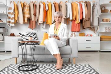 Happy mature female stylist with tablet sitting on sofa against stylish clothes in studio