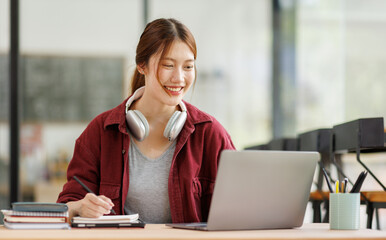 Young asian happy smiling Hispanic Asian student wearing headphones talking on online chat meeting using laptop in university campus or at virtual office. College female student learning remotely.
