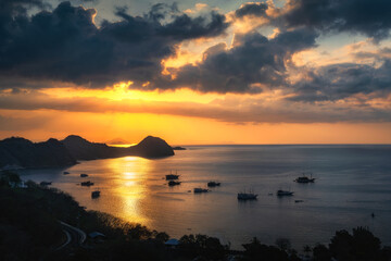 A stunning sunset lights up the horizon, casting golden reflections on the calm sea, with boats...