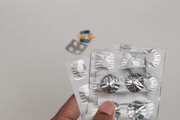 A hand holding several blister packs of medicine with scattered pills in the background,...