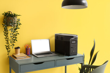 Blank laptop with portable power station on desk near yellow wall in office